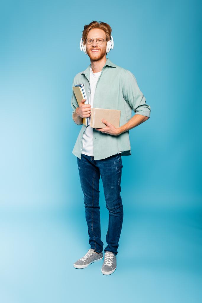 bärtiger Student mit Brille und drahtlosem Kopfhörer, der Bücher und Notizblöcke hält, während er auf blauem Grund steht  - Foto, Bild