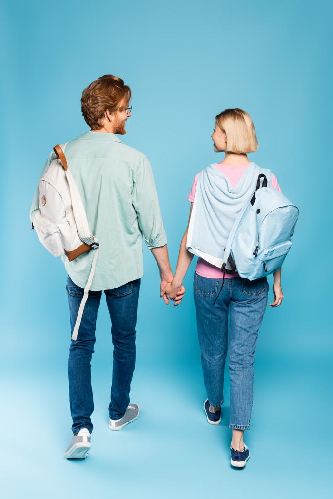 vista trasera de jóvenes estudiantes con mochilas cogidas de la mano mientras caminan sobre azul - Foto, Imagen