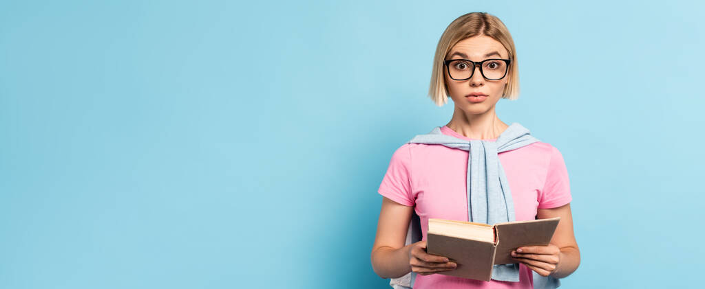 panoramiczna koncepcja blond studentki w okularach trzymającej książkę i odwracającej wzrok na niebiesko - Zdjęcie, obraz