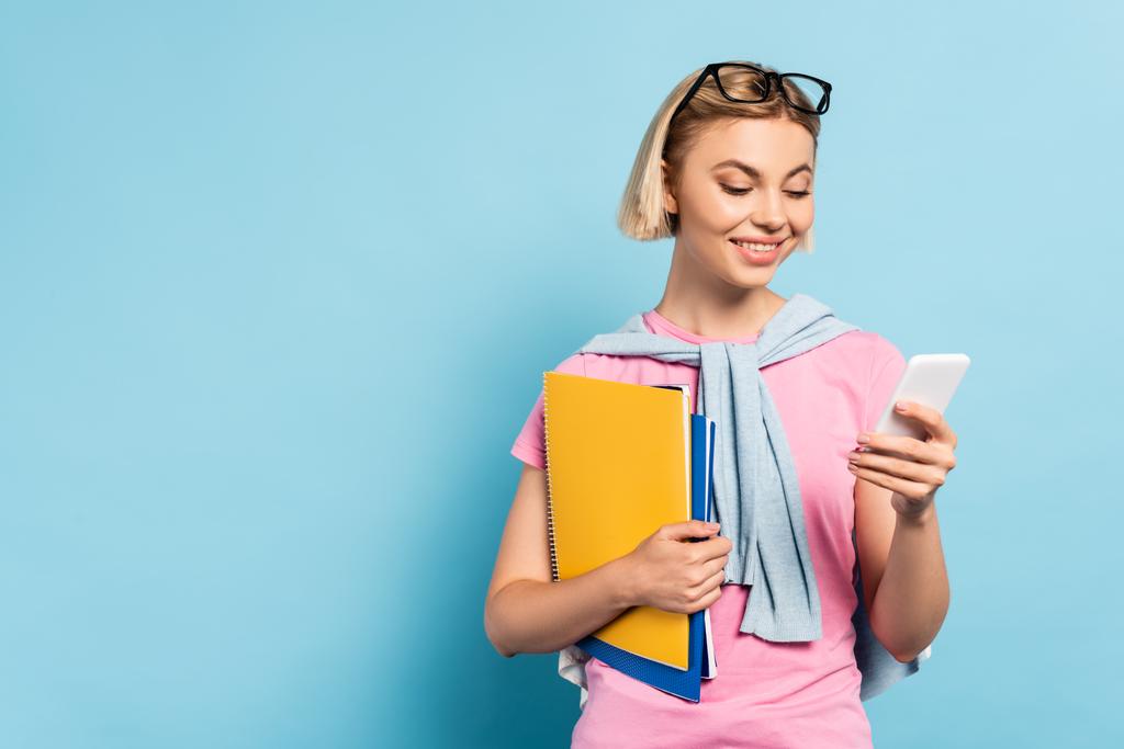 Blonde Studentin hält Notizbücher in der Hand und benutzt Smartphone auf blau - Foto, Bild