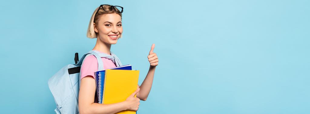 horizontaal beeld van blonde student met rugzak met notebooks en duim omhoog op blauw - Foto, afbeelding