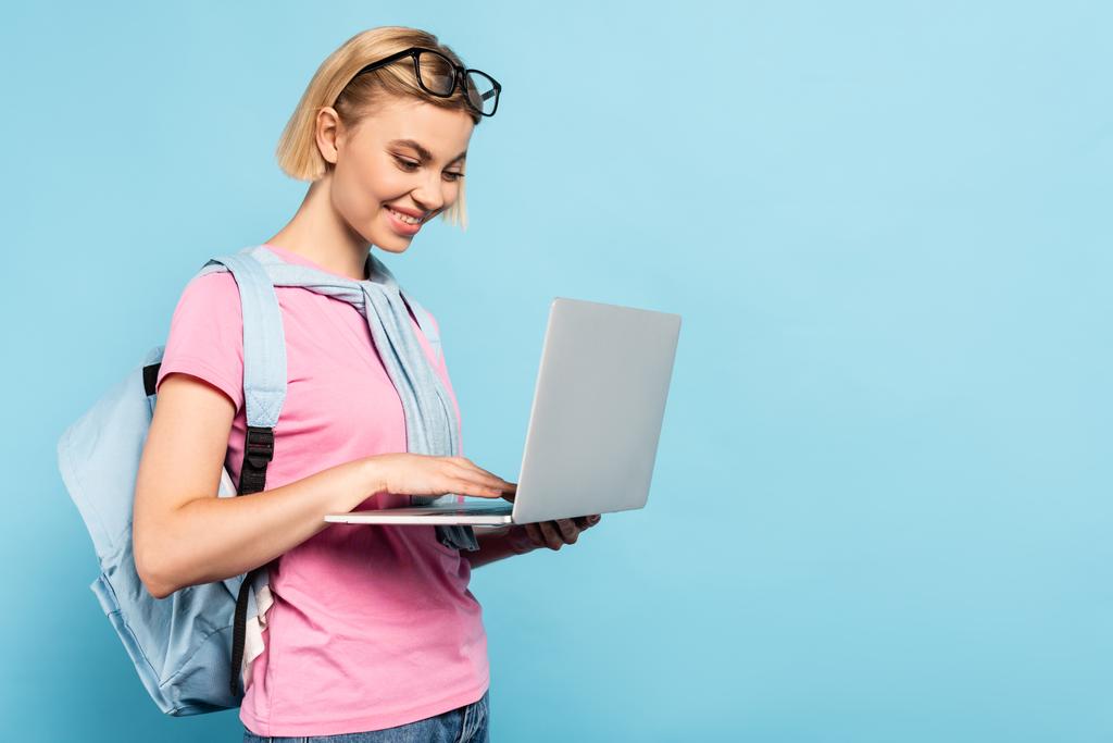 young blonde student with backpack using laptop on blue - Photo, Image
