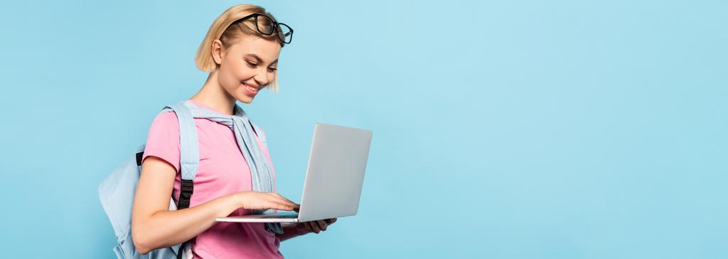 concetto panoramico di giovane studentessa bionda con zaino utilizzando laptop isolato su blu - Foto, immagini