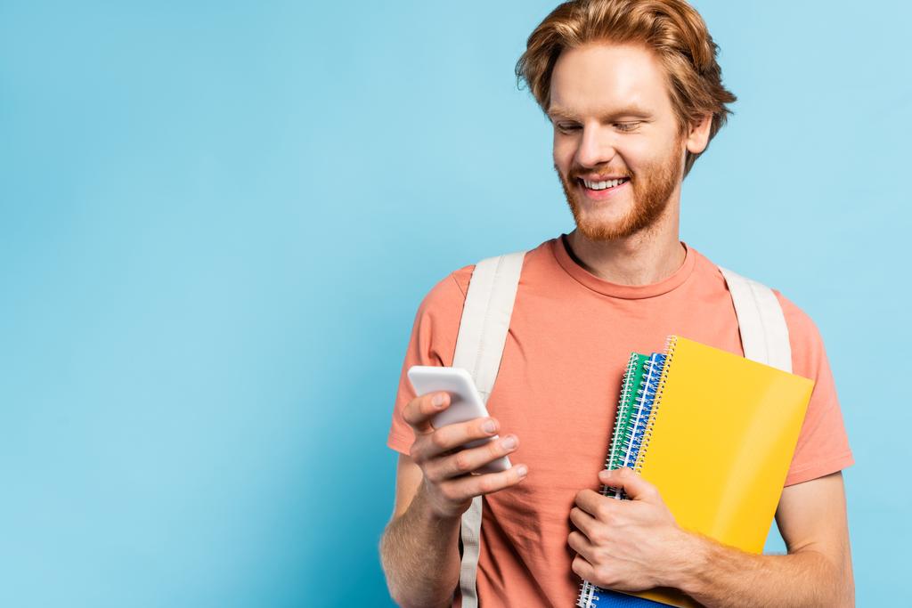 redhead student holding notebooks and using smartphone on blue - Photo, Image