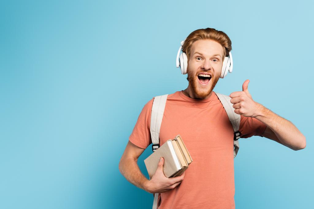 estudiante emocionado en auriculares inalámbricos sosteniendo libros y mostrando el pulgar hacia arriba en azul  - Foto, imagen