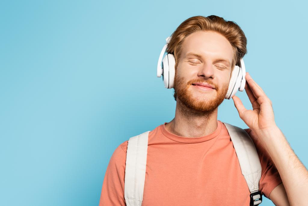 estudante barbudo e ruiva com olhos fechados tocando fones de ouvido sem fio em azul  - Foto, Imagem