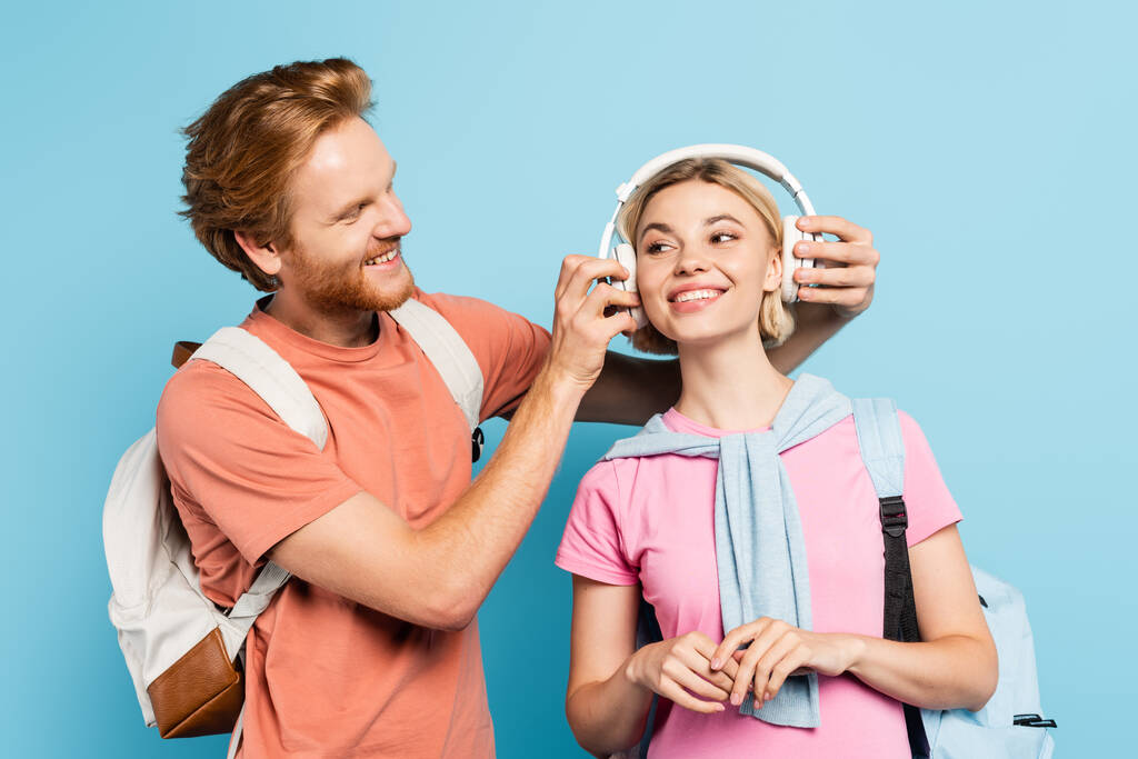 brodaty student z plecakiem dotykający bezprzewodowych słuchawek blond przyjaciela na niebiesko  - Zdjęcie, obraz