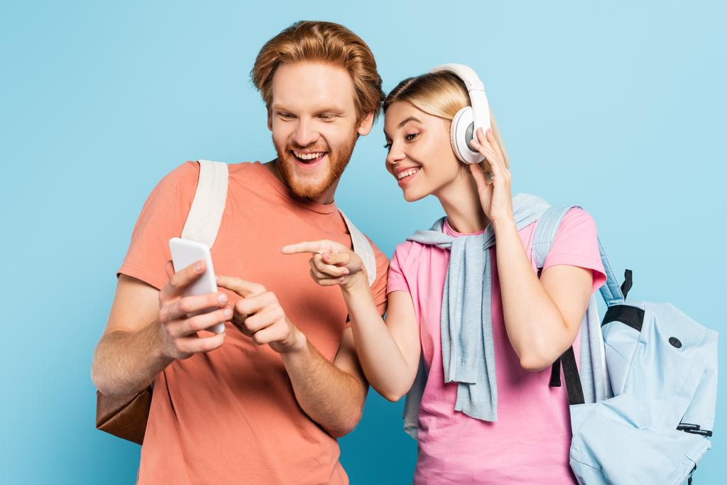 femme blonde écouter de la musique dans les écouteurs sans fil tout en pointant du doigt le smartphone dans les mains de l'étudiant sur bleu - Photo, image