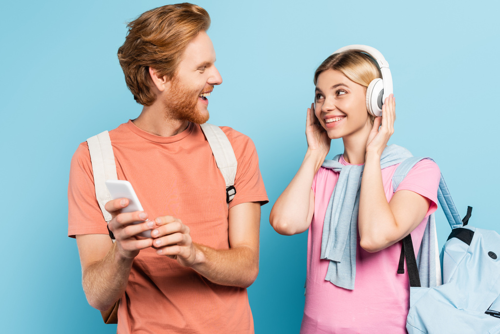 blonde woman listening music in wireless headphones while looking at student with smartphone on blue - Photo, Image