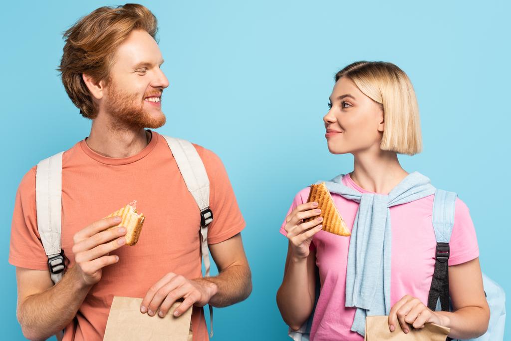 молодые студенты держат бумажные пакеты и сэндвичи, глядя друг на друга в синем - Фото, изображение