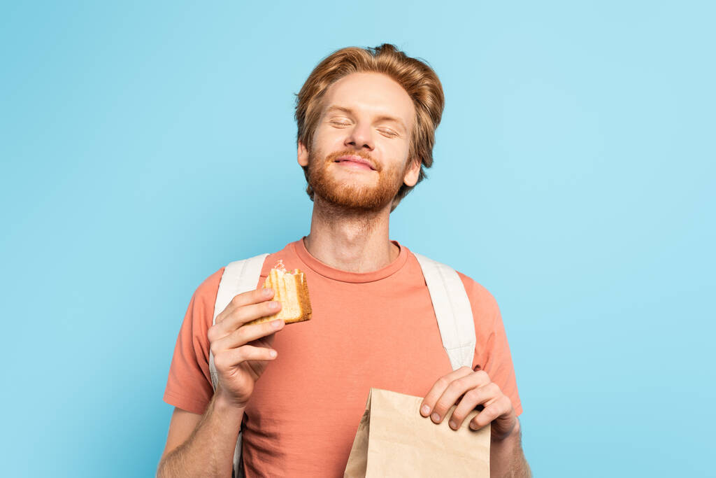 young redhead student with closed eyes holding paper bag and toast bread on blue  - Photo, Image