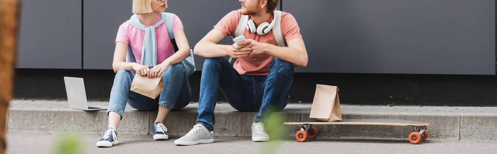 raccolto panoramico di studenti in occhiali seduti vicino skateboard e gadget mentre si guardano fuori - Foto, immagini