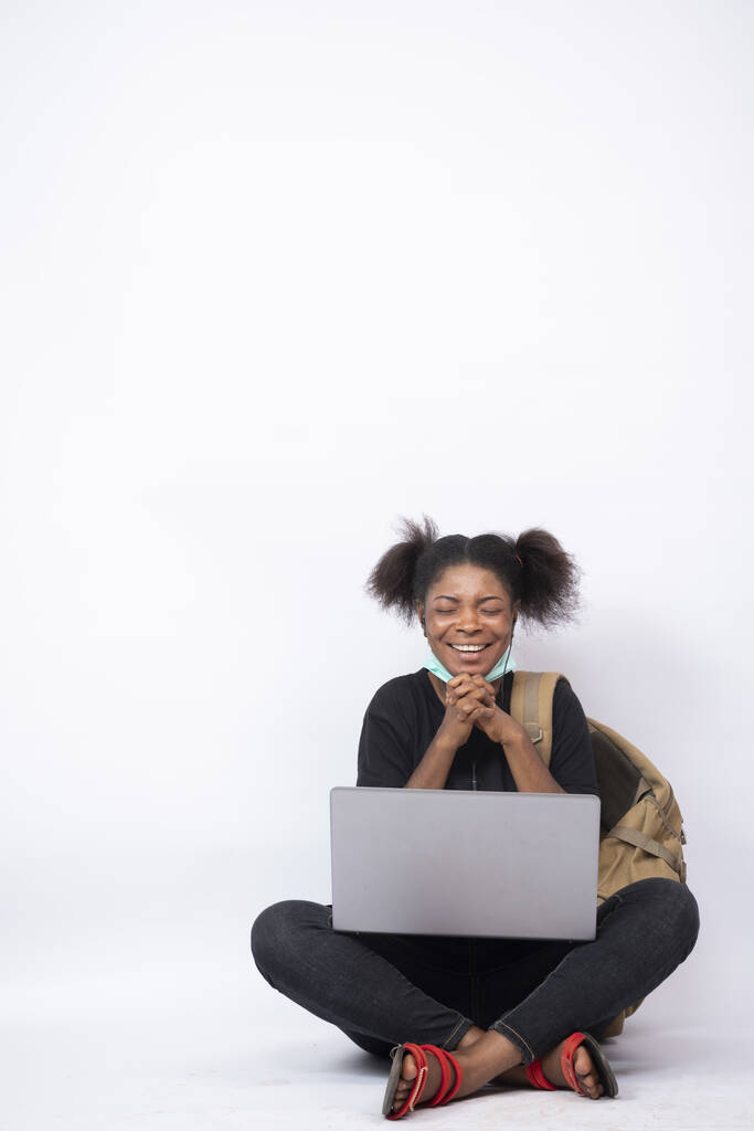 Sırt çantası taşıyan genç bir Afrikalı kadın dizüstü bilgisayar kullanarak bacak bacak bacak üstüne atmış, umutlu. - Fotoğraf, Görsel