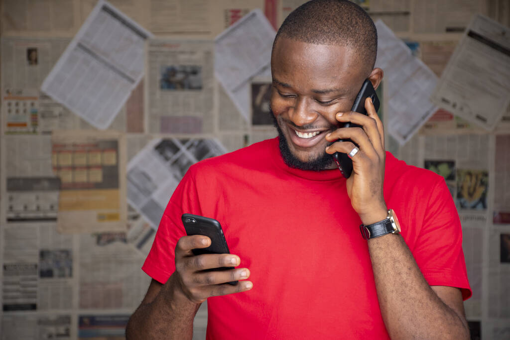 Um jovem homem africano com óculos falando ao telefone enquanto usa outro em uma sala - Foto, Imagem