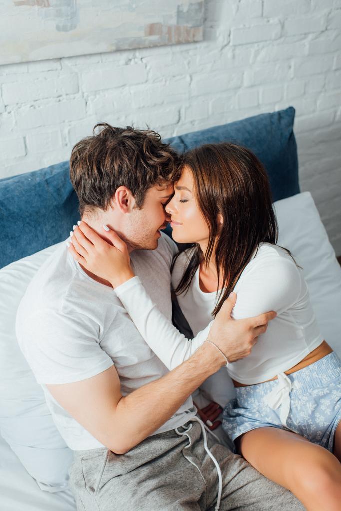 Высокий угол обзора пары в пижаме, сидящей на кровати и трогающей друг друга - Фото, изображение