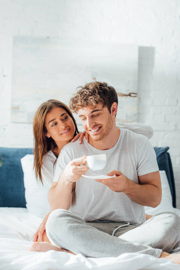 Молодая женщина сидит на кровати и смотрит на парня с чашкой кофе - Фото, изображение