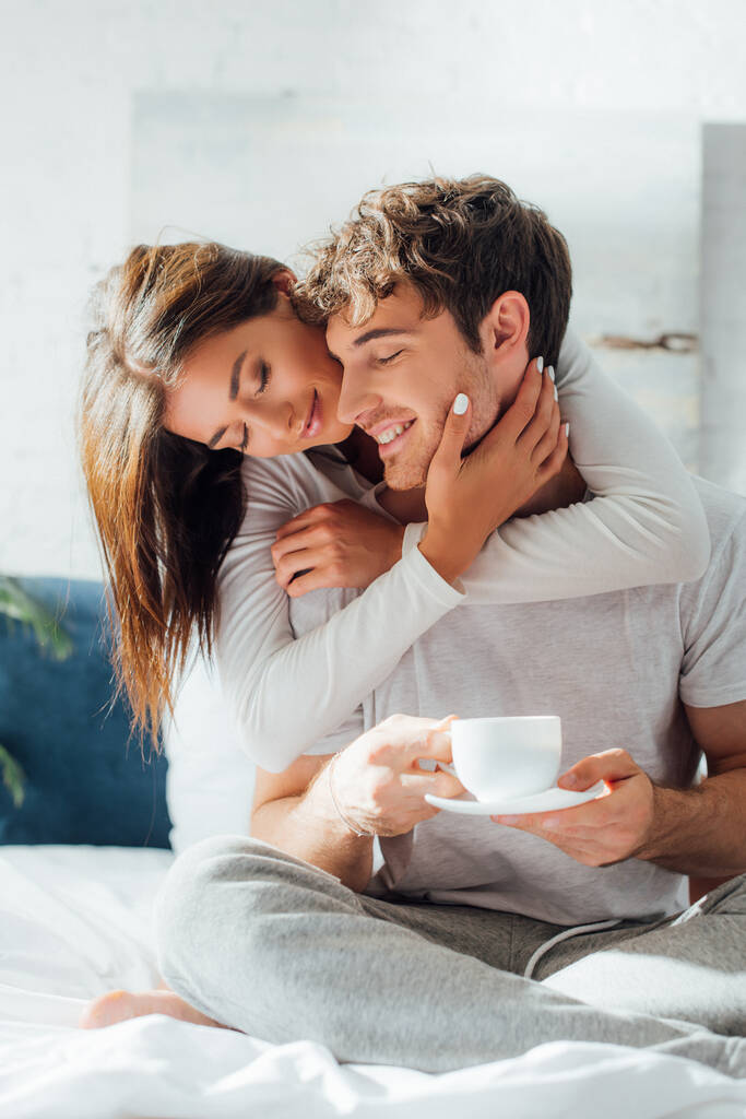 Enfoque selectivo de la mujer joven tocando la cara y abrazando novio con taza de café en la cama - Foto, imagen