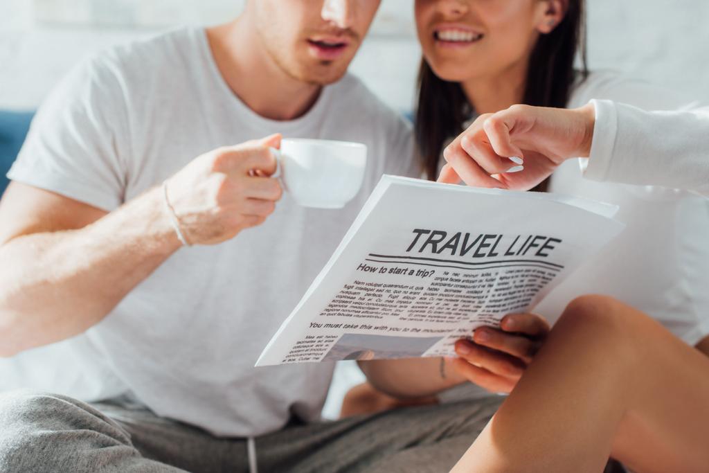 Vue recadrée d'un jeune couple en pyjama tenant un café et lisant un journal avec un article sur la vie de voyage à la maison - Photo, image