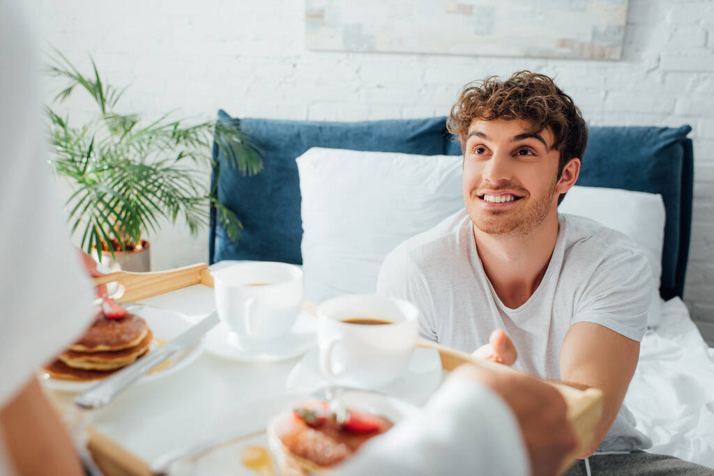 Selective focus of man looking at girlfriend with breakfast and coffee on tray in bedroom  - Photo, Image