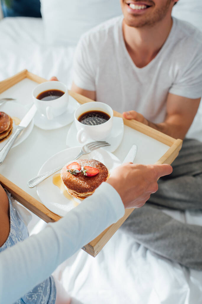 Widok na młodą kobietę trzymającą naleśniki i kawę na tacy śniadaniowej obok chłopaka w piżamie na łóżku  - Zdjęcie, obraz