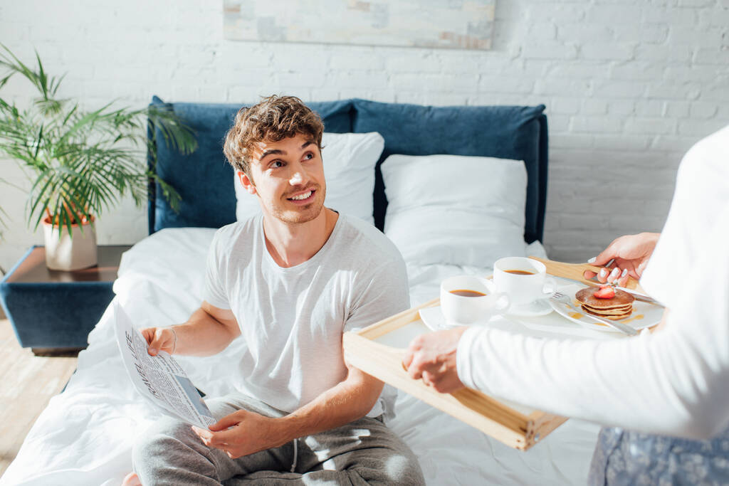 Selective focus of man with newspaper looking at girlfriend holding breakfast on tray in bedroom  - Photo, Image