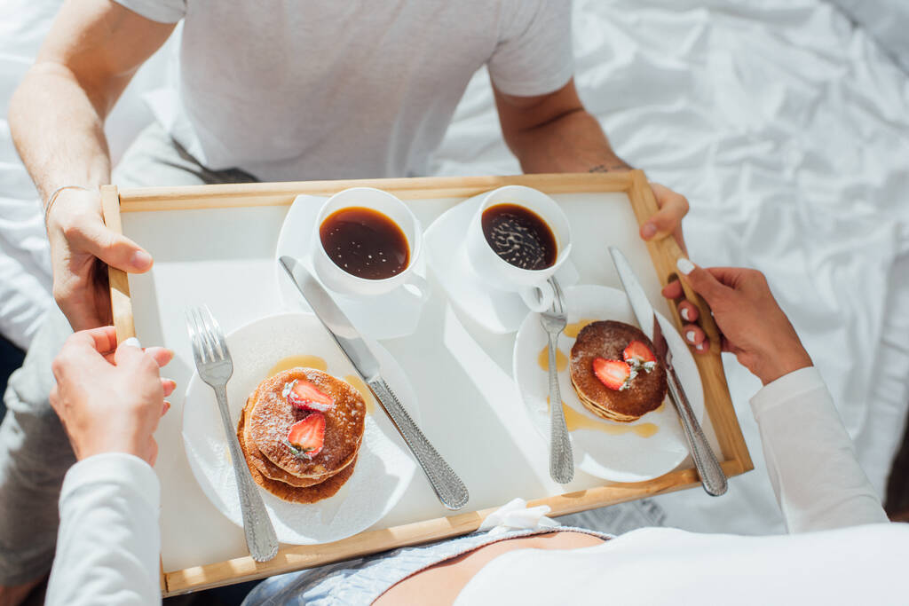 Hochwinkel-Ansicht der Frau mit Pfannkuchen und Tassen Kaffee auf dem Frühstückstablett in der Nähe Freund auf dem Bett  - Foto, Bild