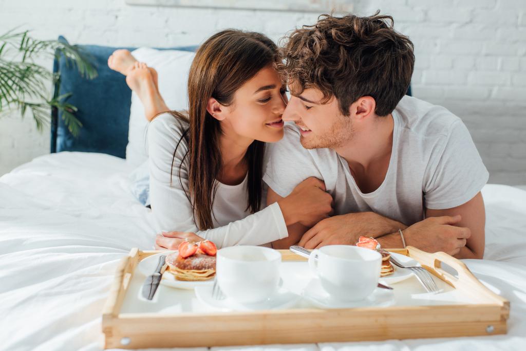 ベッドの上でトレイの上で朝食の近くに横たわる若いカップルの選択的な焦点  - 写真・画像