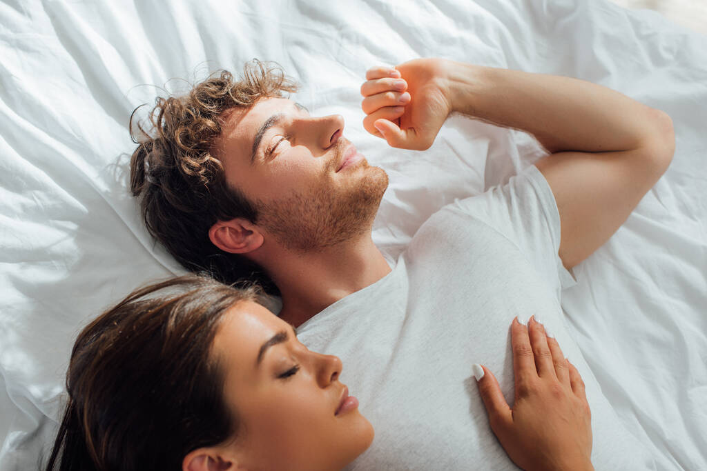 Top view of man stretching while lying on bed near sleeping girlfriend at morning  - Photo, Image