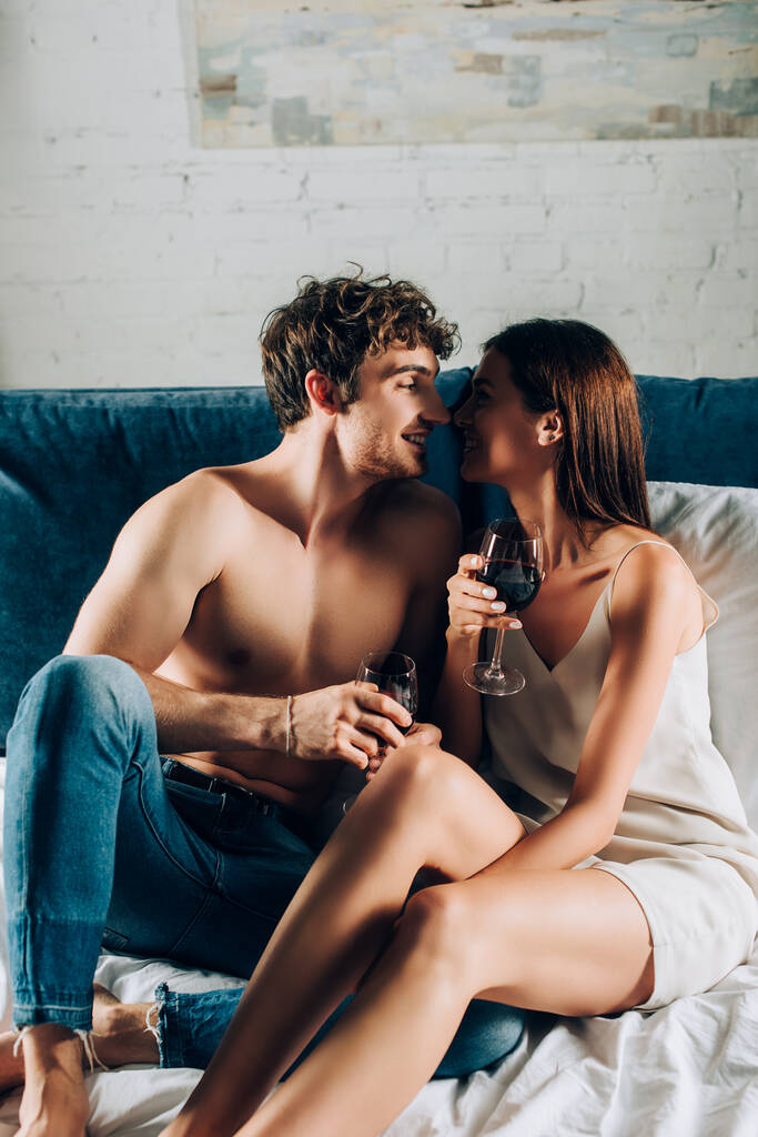 Gespierde man in spijkerbroek met glas wijn in de buurt van vriendin op bed  - Foto, afbeelding
