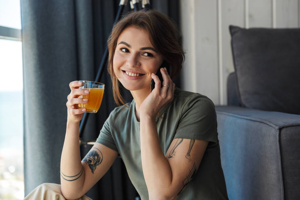 Çekici, gülümseyen genç bir kadın oturma odasının zemininde oturmuş portakal suyu içerken cep telefonu kullanıyor. - Fotoğraf, Görsel