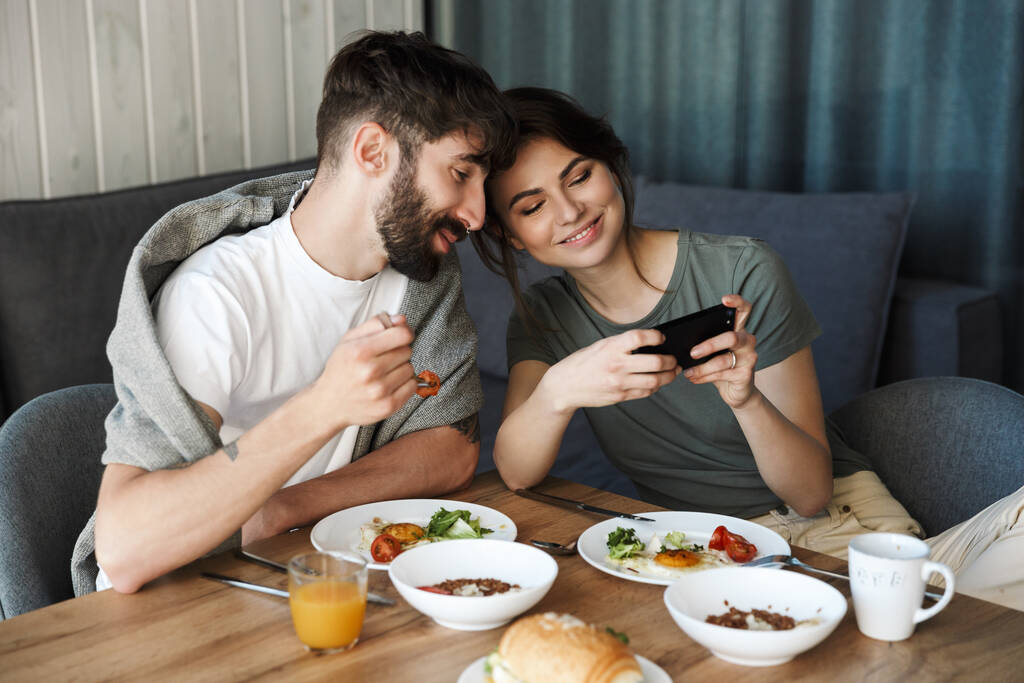 Heerlijk gelukkig jong echtpaar met gezond ontbijt in de keuken, met behulp van mobiele telefoon - Foto, afbeelding
