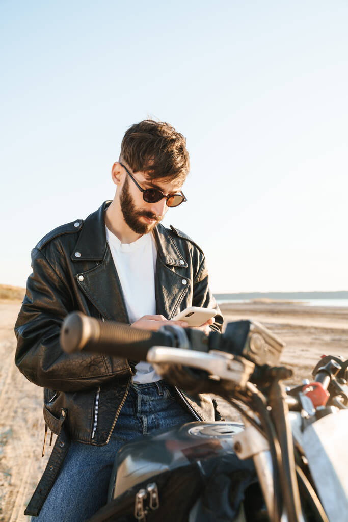 Komea nuori parrakas mies nahkatakissa istuu moottoripyörällä rannalla, käyttäen matkapuhelinta - Valokuva, kuva