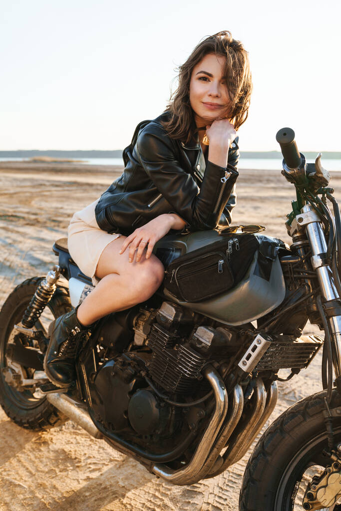 Όμορφη νεαρή γυναίκα που φοράει δερμάτινο μπουφάν κάθεται σε μια μοτοσικλέτα στην παραλία - Φωτογραφία, εικόνα