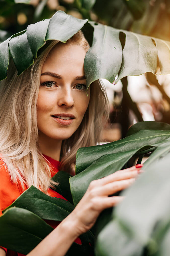 Veselá okouzlující kavkazská blondýnka v červeném tričku, která si užívá času v lese, usmívá se a mezi tropickými listy stojí bílé zuby. Koncept přírody a lidí. - Fotografie, Obrázek