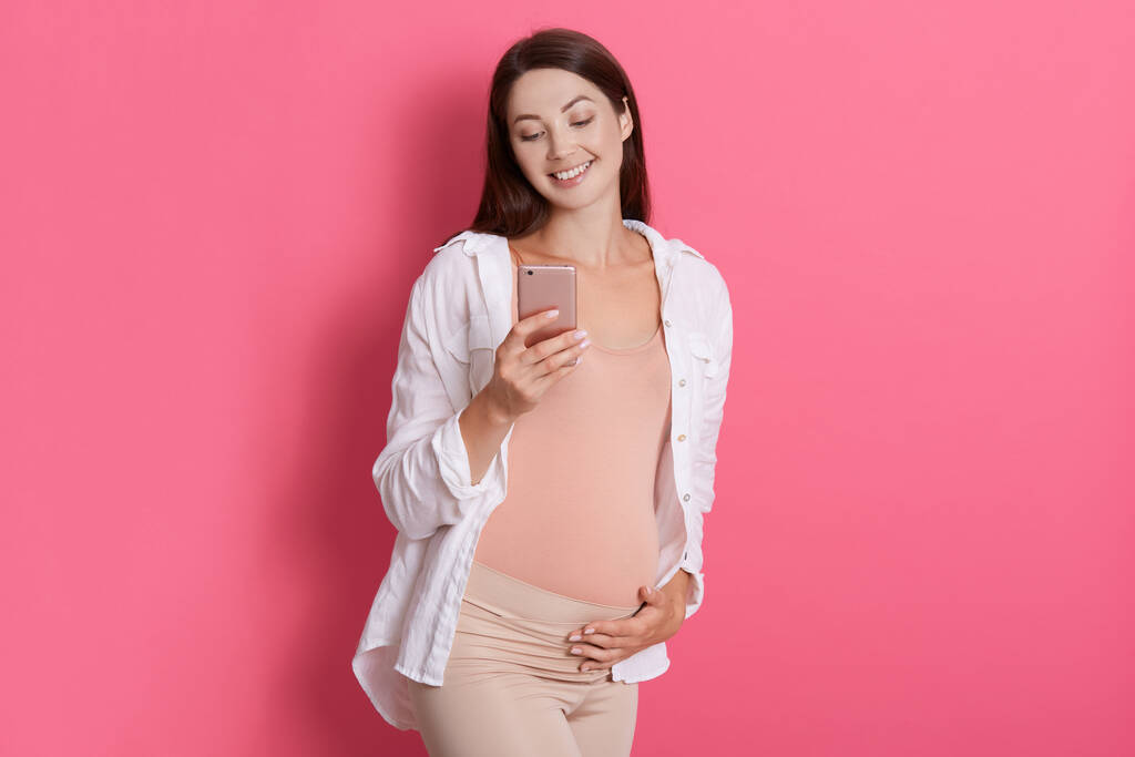 Fröhliche werdende Mutter, die ihr Handy benutzt und mit ihrem Mann plaudert, ihren Schwangerschaftsbauch berührt und glücklich lächelt, süße zukünftige Mama mit Smartphone in der Hand vor rosiger Wand. - Foto, Bild
