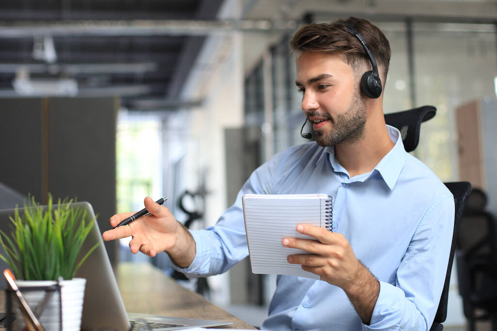 Glimlachende mannelijke callcenter operator met hoofdtelefoon op het moderne kantoor, online informatie raadplegen op een laptop, informatie opzoeken in een bestand om de klant te helpen - Foto, afbeelding