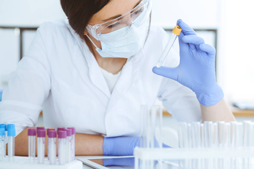 Close-up van professionele vrouwelijke wetenschapper in beschermende brillen die experimenteren met reagentia of bloedonderzoek in het laboratorium. Geneeskunde, biotechnologie en onderzoek. - Foto, afbeelding