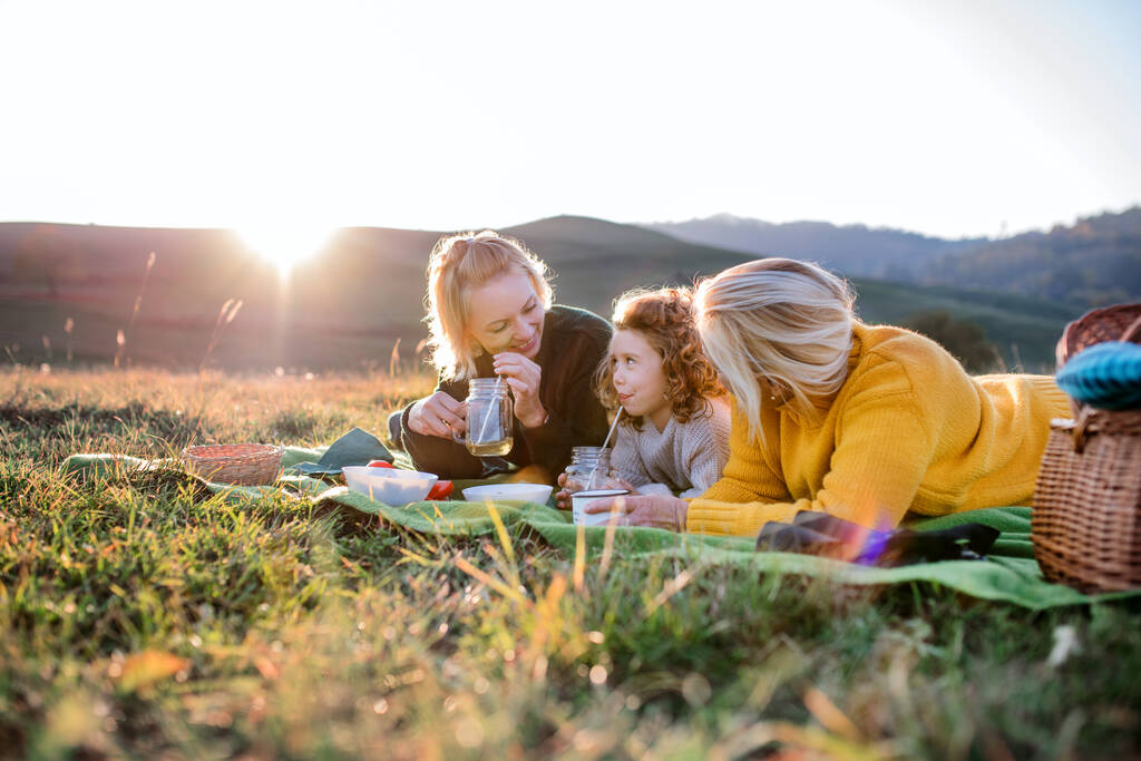 Маленькая девочка с мамой и бабушкой устраивает пикник на природе на закате. - Фото, изображение