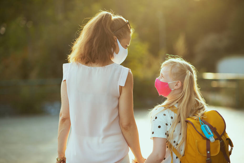 Vida durante a pandemia de covid-19. Visto por trás da jovem mãe e da criança com máscaras, mochila amarela e anti-séptico voltando da escola fora. - Foto, Imagem