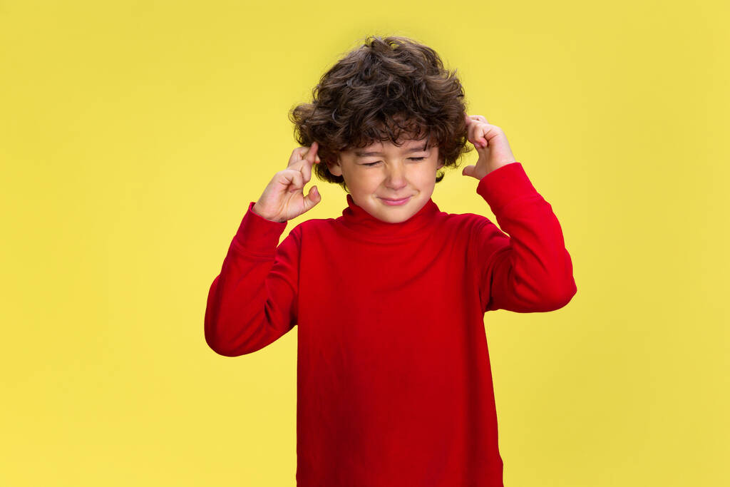 Ένα όμορφο νεαρό σγουρό αγόρι με κόκκινα ρούχα σε κίτρινο φόντο στούντιο. Παιδική ηλικία, έκφραση, διασκέδαση. - Φωτογραφία, εικόνα
