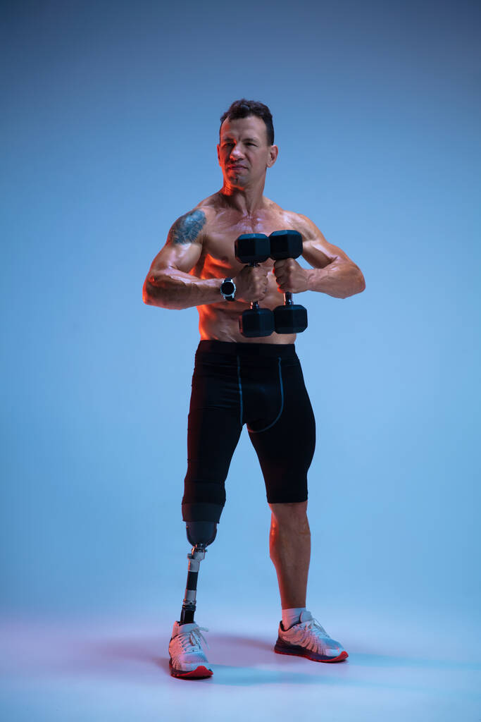 Спортсмен с ограниченными возможностями или ампутация изолированы на синем фоне студии. Профессиональный спортсмен с протезированием ног с грузами в неоновом цвете - Фото, изображение