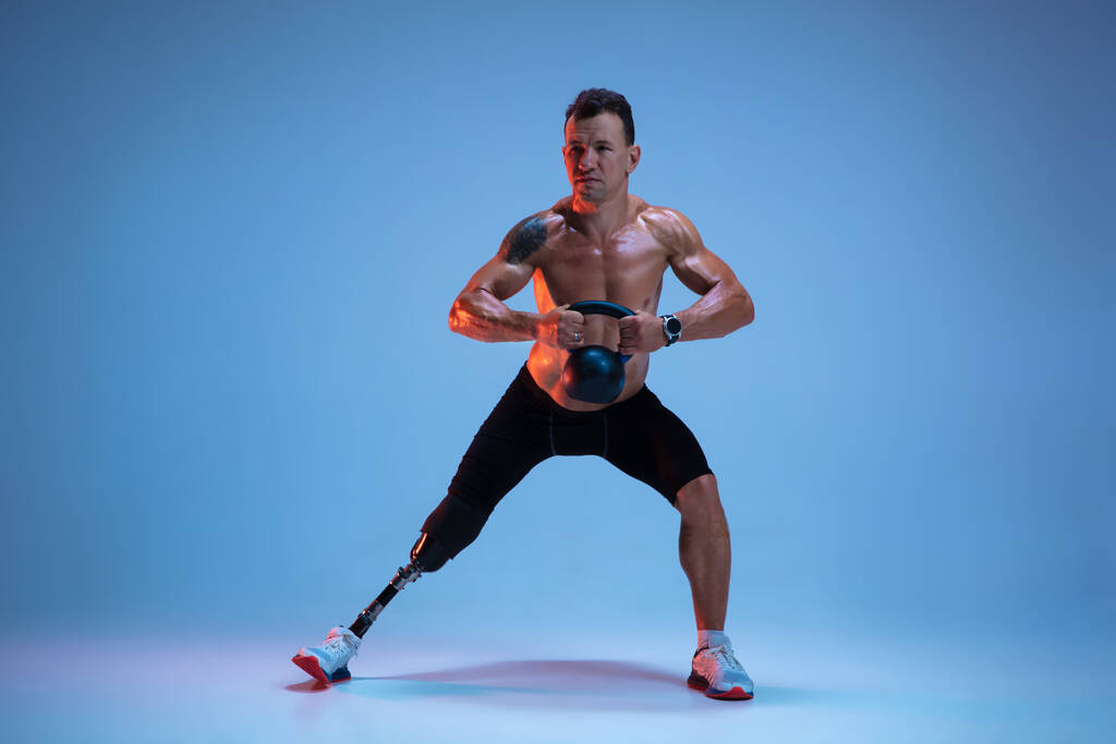 Mozgássérült sportoló vagy amputált, kék stúdió háttérrel elszigetelve. Profi férfi sportoló lábprotézis képzés súlyok neon - Fotó, kép