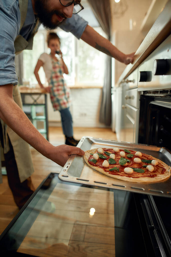 Profesjonalny kucharz robi pizzę w domu. Facet wkłada surowe pizze do nowoczesnego piekarnika do pieczenia. Kobieta stojąca w tle. Hobby, styl życia - Zdjęcie, obraz
