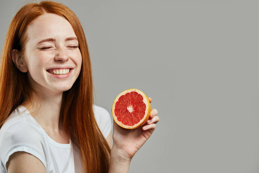 Смеется удивительная очаровательная девушка с закрытыми глазами держит половину грейпфрута - Фото, изображение