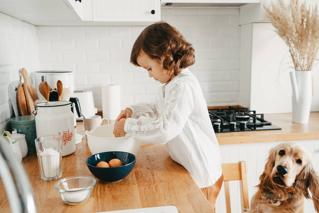 Kleines Mädchen bereitet in der Küche Teig für Pfannkuchen zu. Zusammen mit Spaniel-Hund. Konzept der Nahrungsmittelzubereitung. Lässige Lifestyle-Fotoserie im echten Leben - Foto, Bild