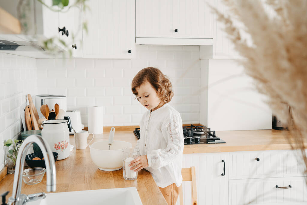 Küçük kız mutfakta krep için hamur hazırlıyor. Yemek hazırlama kavramı, arka planda beyaz mutfak. Gerçek hayatta sıradan bir fotoğraf serisi. - Fotoğraf, Görsel