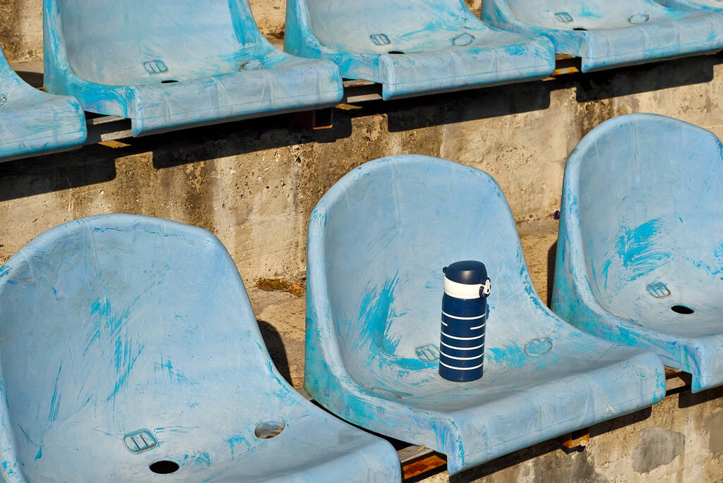 Hay muchos asientos vacíos en el estadio. Botella de agua en el asiento. Bebida para los atletas. Los asientos de plástico están dispuestos en fila. Asientos de ventilador sucios y rayados. - Foto, imagen