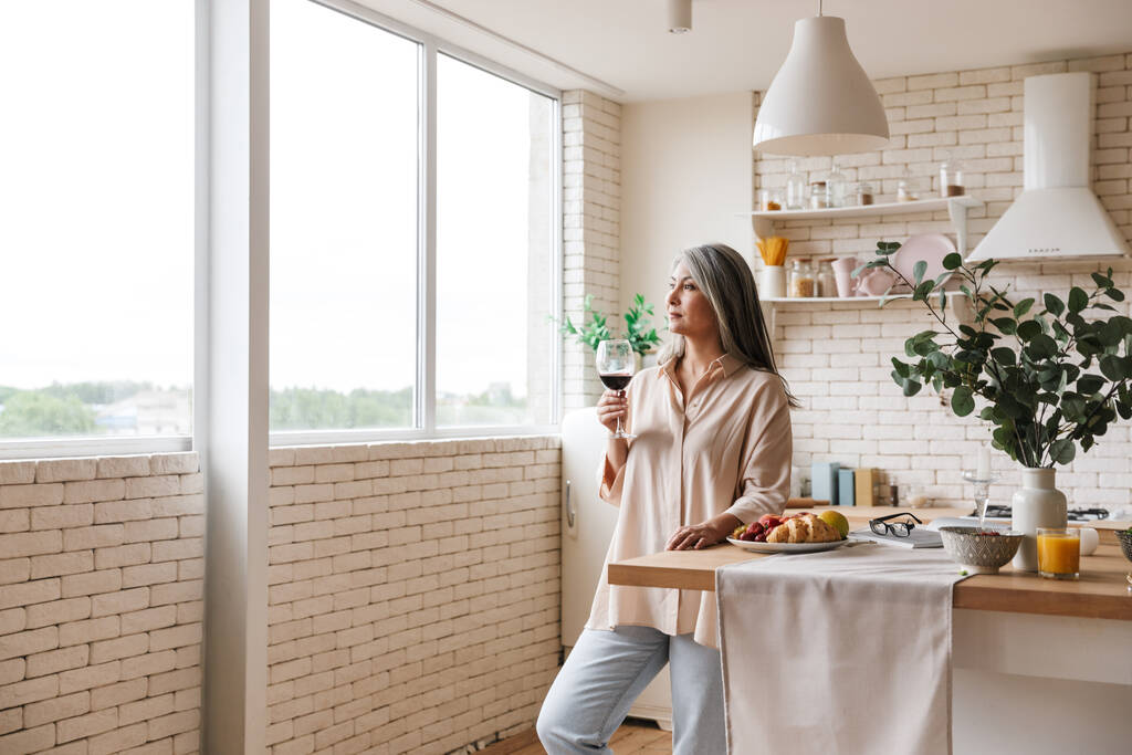 Εικόνα της ώριμης όμορφης γυναίκας που στέκεται στην κουζίνα στο εσωτερικό του σπιτιού πίνοντας κρασί - Φωτογραφία, εικόνα