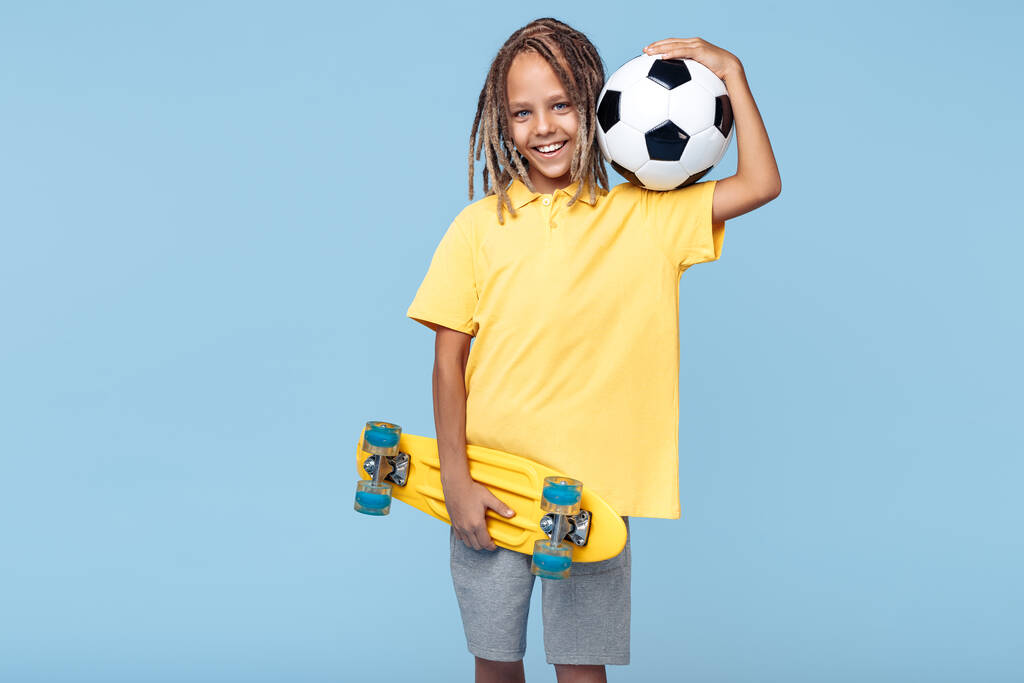 Χαρούμενο μικρό αγόρι με αφρικανικούς φόβους κρατώντας μπάλα ποδοσφαίρου και skateboard πάνω από το μπλε φόντο. - Φωτογραφία, εικόνα