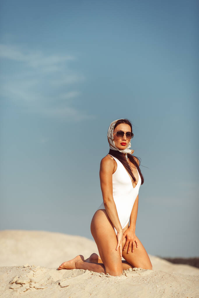 Una joven hermosa chica con una buena figura, alrededor de la arena blanca y el cielo azul, y en ella un traje de baño blanco y hermosos accesorios - Foto, Imagen